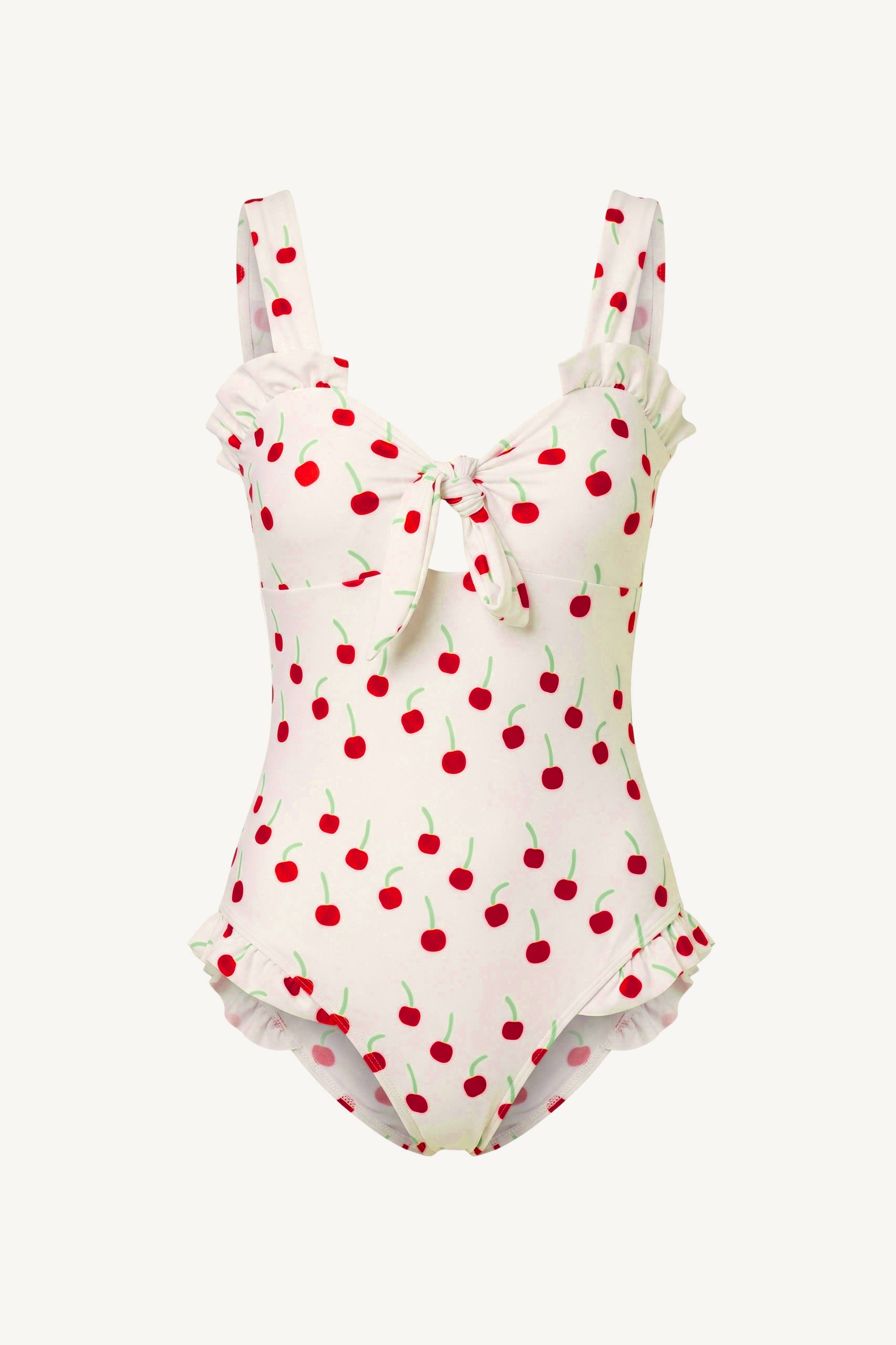 A one-piece swimsuit with a white base and a red cherry print, featuring ruffled straps, a front tie detail, and ruffled trim at the leg openings, shown against a plain off-white background.