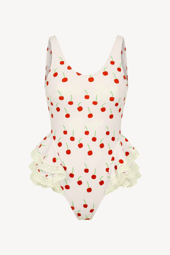 A one-piece swimsuit with a white base, an all-over red cherry print with green stems, and a ruffled white lace hem, shown against a plain off-white background.