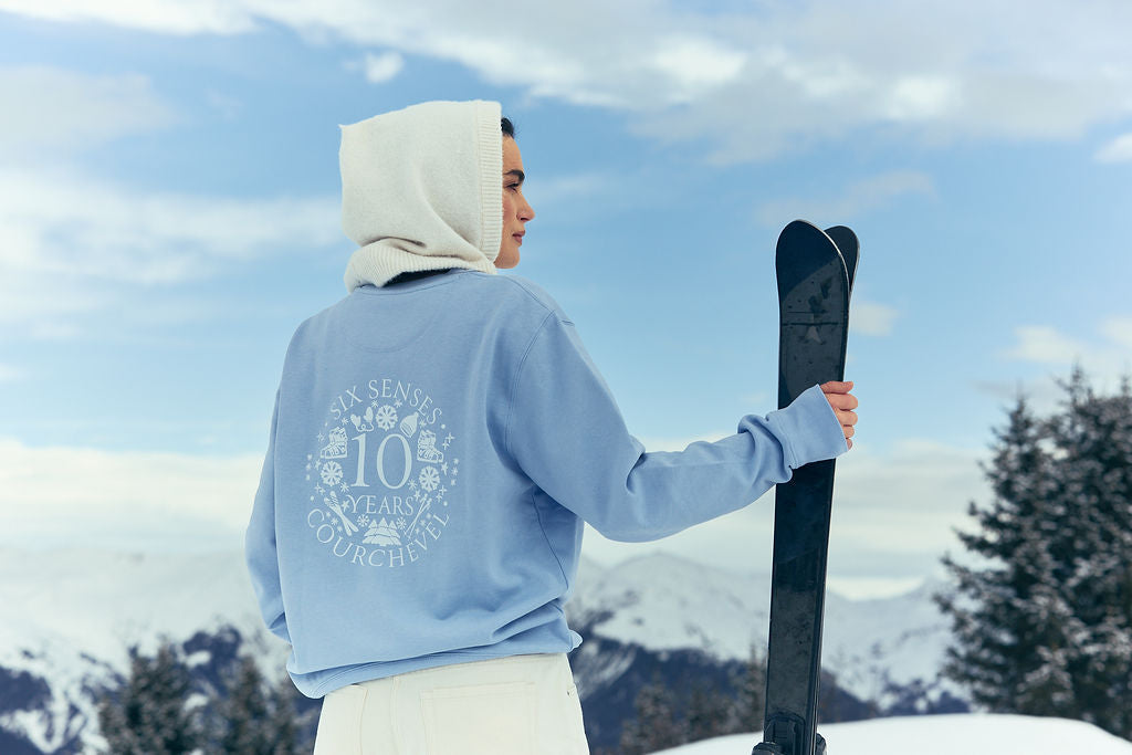 Woman wearing The Courchevel blue sweatshirt standing against snowy mountain holding a pair of skis