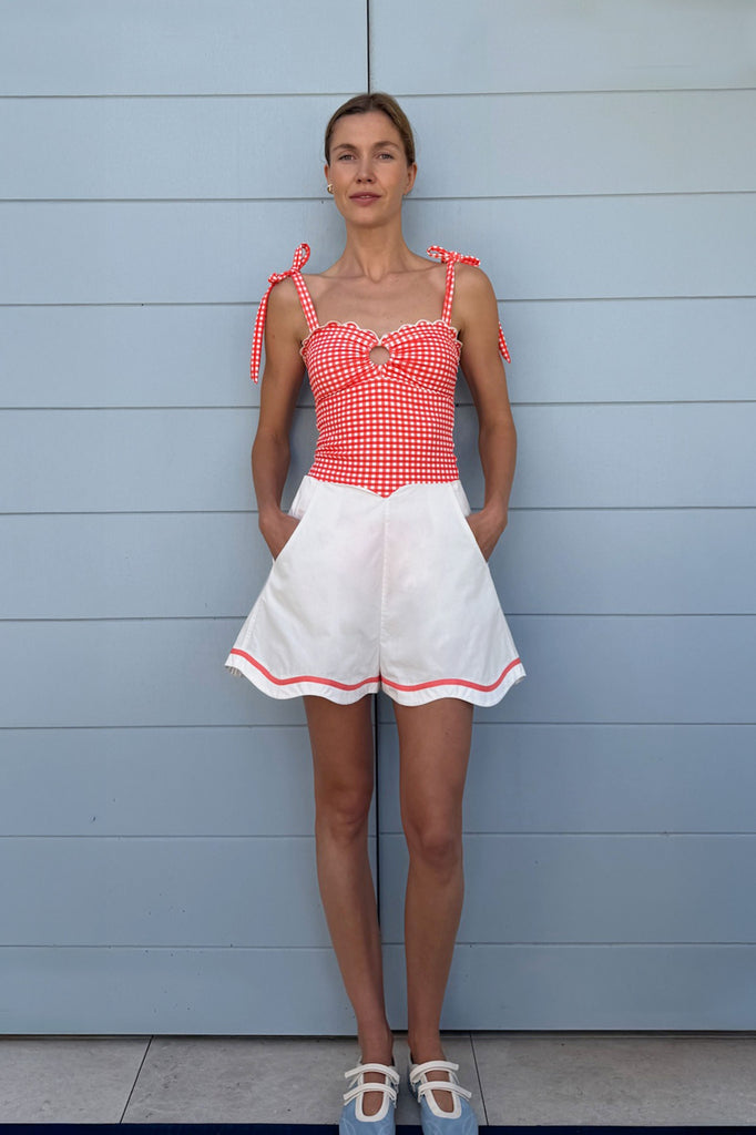 Woman standing against a wall wearing a red and white checkered suit and white shorts with a peach scallop trim. 