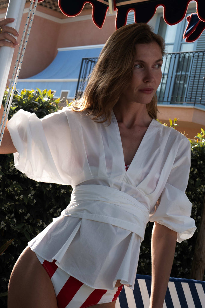 Woman in an ivory wrap top with puffed sleeves and red and white bandeau bathing suit top with matching bottoms standing outdoors.
